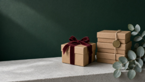 A small gift box tied with burgundy ribbon beside a stack of plain boxes with a blank brass tag, on a frosted-grey ledge against a forest-green wall.
