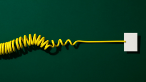 A lemon-yellow coiled telephone cord on a deep emerald-green background, its tangled coils gradually straightening into a single clean line that ends at a blank white paper tag.
